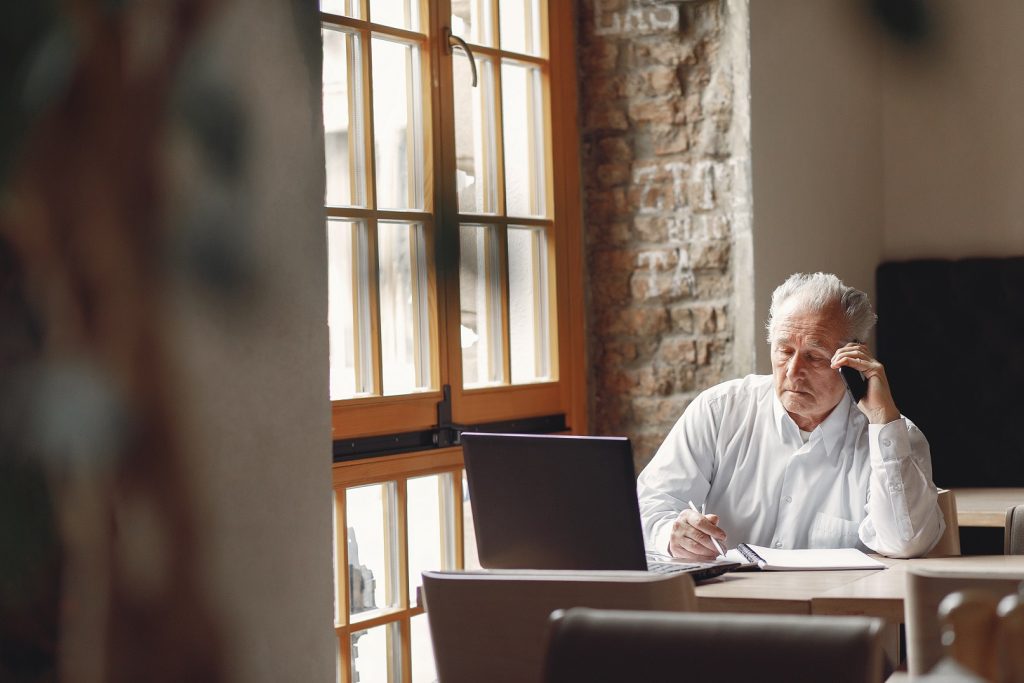 Old Man Sitting At The Table And Working With A Laptop