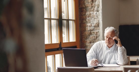 Old Man Sitting At The Table And Working With A Laptop